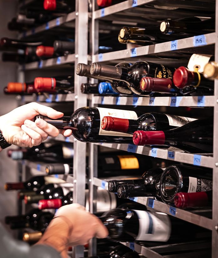 A person carefully selects wine from a well-stocked metal rack with labeled bottles in a restaurant setting.