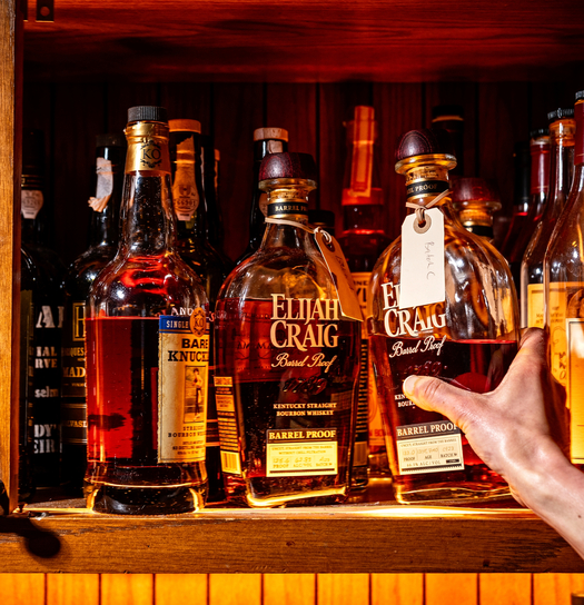 A hand reaches for a bottle of Elijah Craig Barrel Proof whiskey in a wooden shelf displaying various bottles of alcohol.