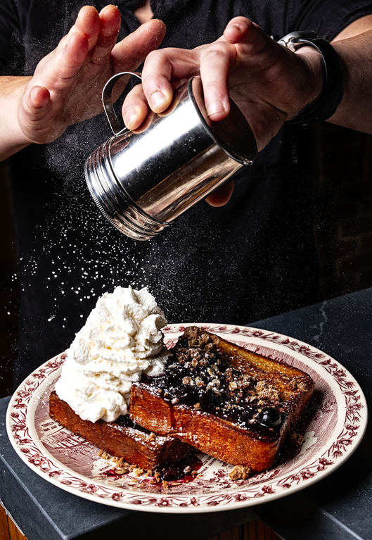 Person sprinkling powdered sugar over a plate of French toast with whipped cream and blueberries.