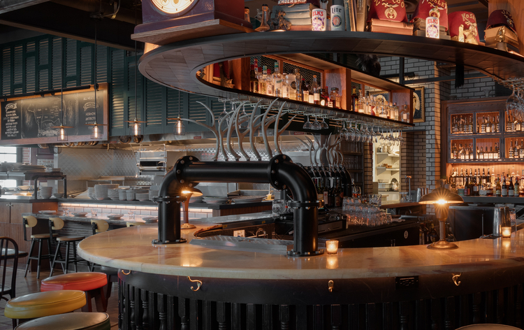 Industrial-style bar with colorful stools, exposed pipes, shelves, and a vintage clock on the back wall.