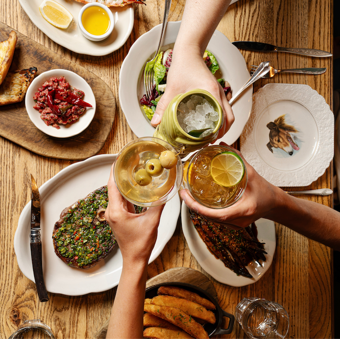 Overhead view of friends toasting cocktails over an Anselm table spread with grilled steak, chimichurri, beef tartare, grilled fish, and fried potatoes